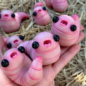 May include: A collection of small, pink, worm-like figurines with smooth, segmented bodies, large glossy black eyes, and small open mouths. The figures have a soft, rounded appearance and are shown in various positions, some resting on a surface.