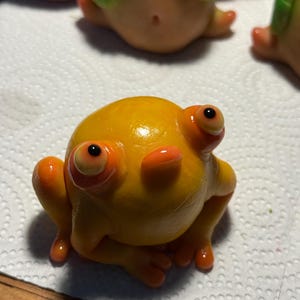 May include: A handmade, glossy, yellow frog figurine with orange detailing. The frog has large eyes and is posed with its legs extended. Other similar figurines are visible in the background. The frog is sitting on a white surface.