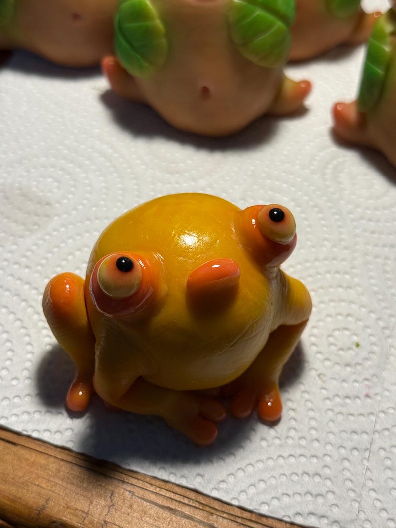 May include: A handmade, glossy, yellow frog figurine with orange detailing. The frog has large eyes and is posed with its legs extended. Other similar figurines are visible in the background. The frog is sitting on a white surface.