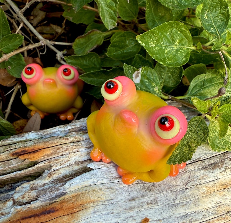 May include: Two quirky frog ornaments with oversized eyes and bright colours. The frogs are mainly yellow with pink and orange details, and red pupils. They are positioned on a weathered wooden surface, surrounded by green leaves.