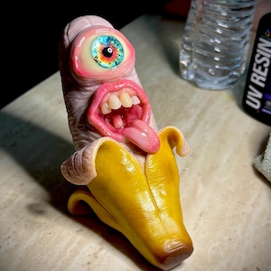 Könnte beinhalten: Eine skulptierte Figur einer geschälten gelben Banane mit einem texturierten, hautfarbenen Oberkörper, der ein einzelnes großes, mehrfarbiges Auge, einen weit geöffneten Mund mit sichtbaren Zähnen und eine herausgestreckte rosa Zunge aufweist.
