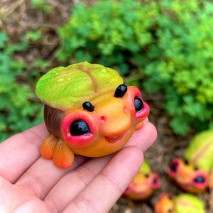 May include: A whimsical, handmade figurine of a smiling, cartoon-like creature. The figurine features a yellow and orange face with large, black eyes and a green leaf-like head covering. The creature has small orange feet and a brown body. Several similar figurines are visible.