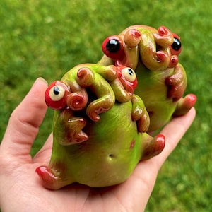 May include: Two quirky, handmade sculptures of green and red creatures with large eyes and tentacle-like features. The figures are held in a hand against a blurred green backdrop, highlighting the unique, artistic design.