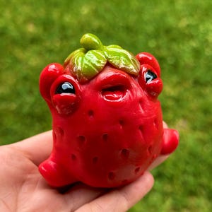 May include: A quirky, red strawberry-shaped sculpture with black eyes, a small mouth, and green leaves. The sculpture has a textured surface with small, dark holes, and small red appendages. The sculpture is held in a hand against a blurred green background.