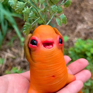 May include: A whimsical orange sculpture resembling a creature with large black eyes, a small open mouth, and a green leafy sprout. The sculpture is held in a hand, with a blurred background of greenery.