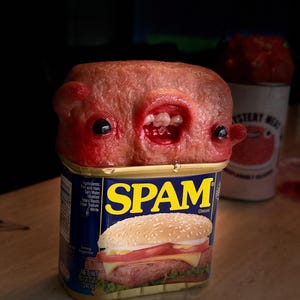 Puede incluir: Una escultura surrealista de una lata de Spam con una criatura grotesca y carnosa que emerge de la parte superior. La lata está etiquetada como "SPAM Classic" y presenta una ilustración de hamburguesa. Una segunda lata, "MYSTERY MEAT", está en el fondo.