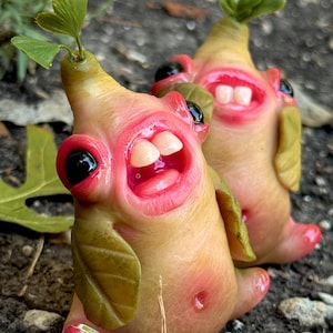 May include: Two whimsical, handmade sculptures of smiling, humanoid creatures. They have large, black eyes, pink lips, and visible teeth. The creatures are tan with pink accents and green leaf-like appendages. The sculptures are set against a natural outdoor background.