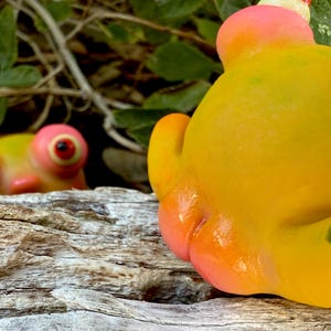 May include: A close-up of a yellow and pink frog ornament with large, red-centered eyes. The figurine's textured surface and rounded features are in focus. Another similar ornament is visible in the background, partially obscured by foliage.