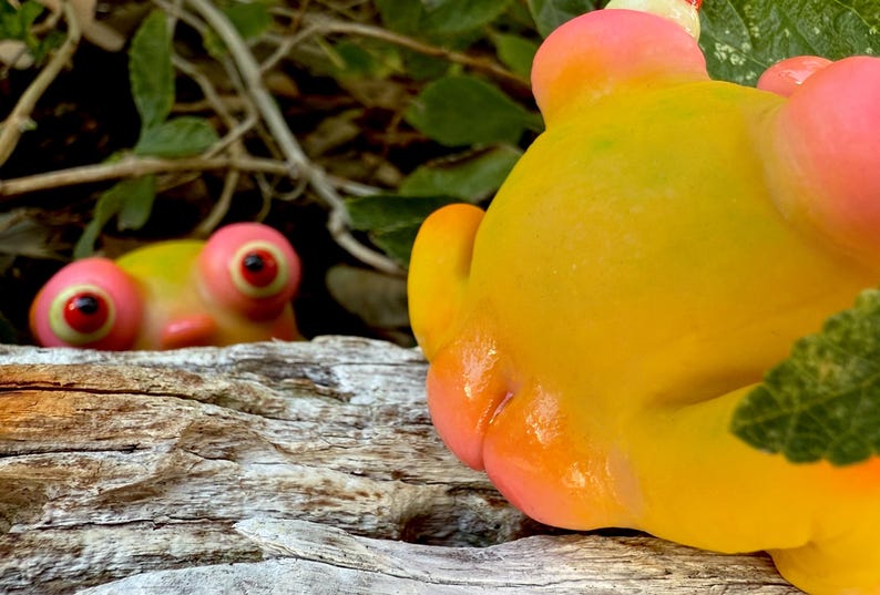 May include: A close-up of a yellow and pink frog ornament with large, red-centered eyes. The figurine's textured surface and rounded features are in focus. Another similar ornament is visible in the background, partially obscured by foliage.