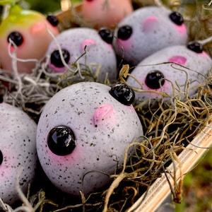 May include: A collection of whimsical, speckled, grey and pink egg-shaped ornaments with black eyes and pink ears, nestled in a bed of dried moss. Some ornaments are light green and pink. The ornaments are in a wooden container.