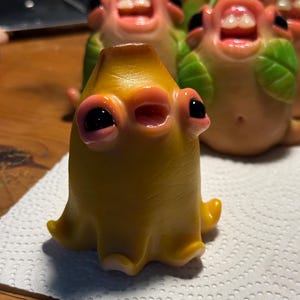 May include: A group of quirky, handmade figurines. The front figure is yellow with large black eyes and a pink mouth. Other similar green and pink creatures are in the background. The figurines have a cartoonish, playful design.