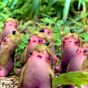 May include: A group of handmade sculptures of fantastical creatures. They have pinkish-purple bodies, large black eyes, and leaf-like head coverings with black accents. The figures are set amongst green foliage, creating a natural scene.