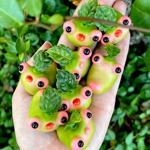 May include: A collection of small, green frog figurines with pink accents and black eyes. The frogs are made of polymer clay and have a whimsical, cartoonish style.