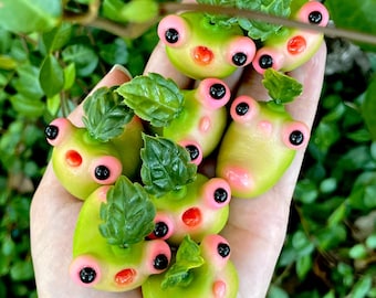 Puede incluir: Una colección de pequeñas figuritas de rana verde con detalles rosas y ojos negros. Las ranas están hechas de arcilla polimérica y tienen un estilo caprichoso y de dibujos animados.