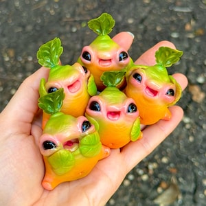 May include: Five smiling figurines resembling radishes, held in a hand. They have green leafy tops, pink faces, and large black eyes. The bodies are a gradient of yellow and orange. A charming, handcrafted design.