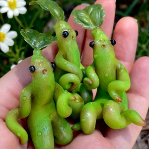 May include: Three small green hand-crafted figurines with leaf-like tops and black eyes. The figurines are made to look like small, whimsical creatures.