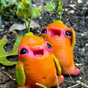 May include: Two quirky orange sculptures with pink mouths and black eyes, reminiscent of cartoon characters. Green leafy sprouts grow from the top of their heads. The figures have small arms and feet, set against a natural, earthy background.