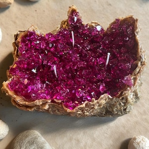May include: A geode-shaped candle featuring a textured, metallic gold exterior and a center filled with vibrant, translucent magenta crystal-like fragments. The candle includes three white wicks embedded within the jagged, crystalline surface.
