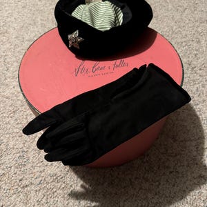 May include: A black velvet hat with a jeweled pin and a green and white striped lining. The hat is sitting on a pink box with the text "Stix, Baer & Fuller, Saint Louis" printed on it. A pair of black gloves are also on the box.