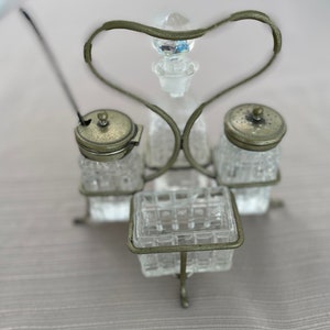 May include: A silver metal condiment holder with a heart-shaped design. The holder has four glass containers with silver lids and a central glass container with a silver stopper. The holder is on a light brown surface.
