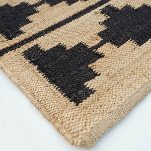 May include: Close-up of a natural fiber rug with a geometric pattern in black and beige. The rug has a woven texture and a fringed edge.