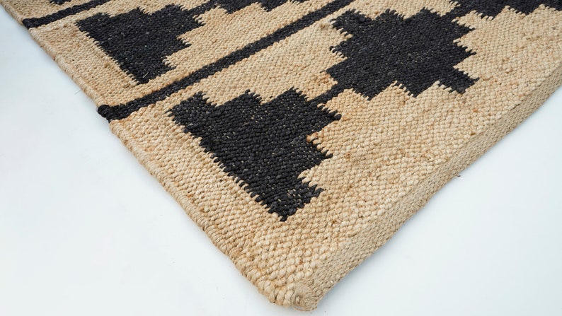 May include: Close-up of a natural fiber rug with a geometric pattern in black and beige. The rug has a woven texture and a fringed edge.