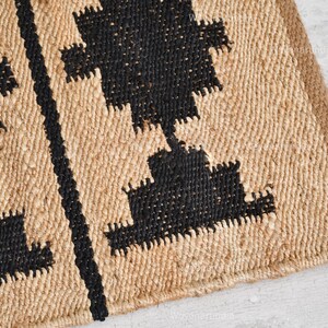 May include: A close-up of a woven jute rug with a geometric pattern in black. The rug has a natural, earthy look and feel.