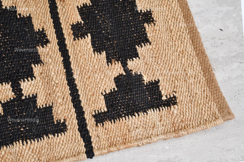 May include: A close-up of a woven jute rug with a geometric pattern in black. The rug has a natural, earthy look and feel.