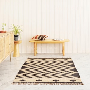 May include: A rectangular rug with a geometric zigzag pattern in black and beige. The rug has a fringed edge and is placed on a light-coloured floor. A wooden bench and cabinet are in the background.