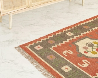 May include: A rectangular rug with a terracotta background and a geometric pattern. The rug has a zigzag border in grey and green, and a central design with floral motifs. The rug has a fringed edge. Measurements are in centimetres.