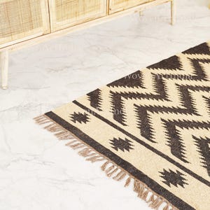 Op de afbeelding: Een beige en bruin geometrisch patroon tapijt met een zigzag ontwerp. Het tapijt heeft een rand met stervormige vormen en franjes langs één rand. Het tapijt is gemaakt van een geweven materiaal.