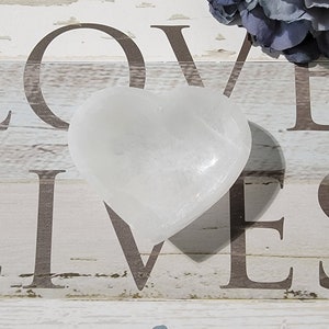 May include: A heart-shaped, white selenite crystal. The crystal is smooth and polished, reflecting light. The heart sits on a wooden surface with the word "LOVE" printed in brown. A blue flower is in the background.