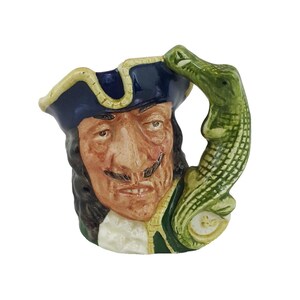 May include: A ceramic mug in the shape of a pirate's head, with a detailed face and a tricorn hat. The handle is a green alligator. The mug is painted in shades of brown, blue, green, and cream, with a detailed design.
