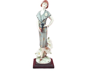 Könnte beinhalten: Vintage Porzellanfigur einer Frau in einem Kleid, Hut und Schuhen im Flapper-Stil, die eine Handtasche hält. Die Figur ist hellblau mit floralen Akzenten und steht auf einem Sockel mit weißen Blumen.