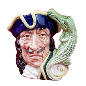 May include: A ceramic mug with a detailed, three-dimensional design of a man's face with a mustache and a tricorn hat. A green alligator is sculpted on the side. The mug has a cream, blue, and green colour scheme.