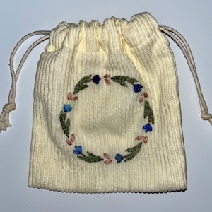 May include: A small, off-white, corduroy drawstring bag with a floral embroidery design. The embroidery is a wreath of green leaves, blue and pink flowers.