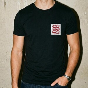 May include: Black crew neck t-shirt with a small square graphic on the chest. The graphic shows a series of red lip imprints. The short-sleeved shirt is likely made of a soft fabric.