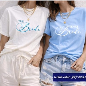 Op de afbeelding: Twee T-shirts, één witte en één hemelsblauwe, met de woorden "The Bride" en "Team Bride" erop gedrukt. Het hemelsblauwe shirt wordt gecombineerd met een spijkerbroekshort. Het witte shirt wordt gecombineerd met een witte broek. Beide shirts zijn korte mouwen.