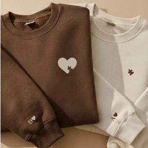 Op de afbeelding: Een donkerbruin sweatshirt met een wit hart met een puzzelstukje en een crèmekleurig sweatshirt met een bruin puzzelstukje. De bruine mouw heeft de tekst D gevolgd door een hartje, en de crèmekleurige mouw heeft de tekst A gevolgd door een hartje.