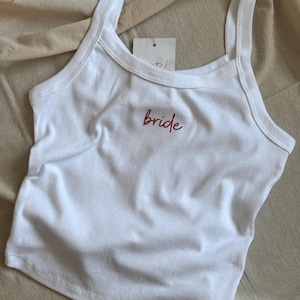 Può includere: Canotta bianca con spalline sottili e la parola "bride" ricamata in rosso. La parte superiore è realizzata in un materiale morbido e ha un'etichetta attaccata. Un capo casual per un matrimonio.