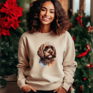 Könnte beinhalten: Ein beiger Pullover mit einer Aquarell-Illustration eines braunen und schwarzen Hundes mit lockigem Fell. Der Hund schaut nach rechts im Bild.