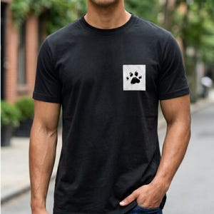 May include: Black t-shirt with a white square pocket displaying a black paw print. The shirt has a crew neck and is made of a soft material. The model is wearing blue jeans.