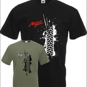 Pode incluir: Duas camisetas com um gráfico de banda de rodagem de pneu em preto e branco e o texto "Africa Twin" em vermelho. A camiseta à esquerda é verde oliva e a camiseta à direita é preta.