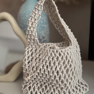 Puede incluir: Una pequeña bolsa de crochet de color blanco roto con un asa anudada. La bolsa está hecha de un material grueso parecido a una cuerda y tiene un tejido suelto y abierto.