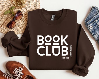 May include: A brown sweatshirt with the text "BOOK CLUB" in white letters. The text is in a bold, blocky font. Below the text is "CUSTOM NAME" and "EST. 2023" in smaller white letters.
