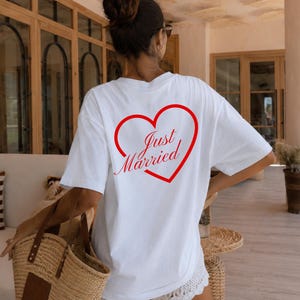 Op de afbeelding: Wit T-shirt met een rode hartafbeelding en de tekst "Just Married". Dit casual shirt is ideaal voor pasgetrouwden. Het model draagt een witte short en een strooien draagtas. De setting is buiten.