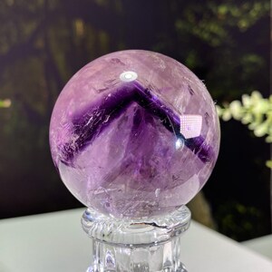 Puede incluir: Una gran esfera de cristal de amatista púrpura sobre un soporte de vidrio transparente. La esfera tiene un color púrpura intenso y está pulida con un acabado liso.