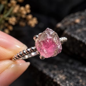 Könnte beinhalten: Ein Silberring mit einem rohen, ungeschliffenen rosa Turmalin-Edelstein in einer Vier-Krappen-Fassung. Die Ringschiene hat eine gedrehte, seilartige Textur, die für ein detailliertes metallisches Finish sorgt, das mit der unregelmäßigen Form des Steins kontrastiert.