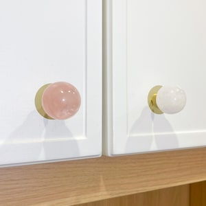 May include: Two cabinet knobs with a gold base and a round, polished stone top. One knob is pink and the other is white.
