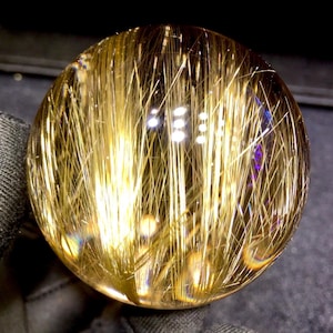 Puede incluir: Una bola de cristal transparente con inclusiones doradas en forma de aguja. Las inclusiones crean un efecto visual llamativo, que se asemeja a hebras de oro. El cristal refleja la luz, creando una exhibici&oacute;n brillante e iridiscente. Un art&iacute;culo de decoraci&oacute;n &uacute;nico.